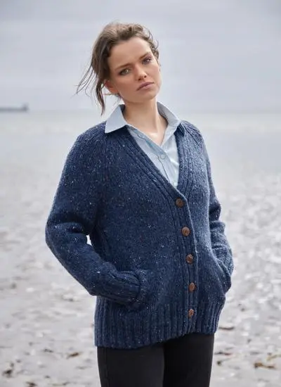 A woman with brown hair wearing a blue wool cardigan is standing on a beach with the ocean behind her.